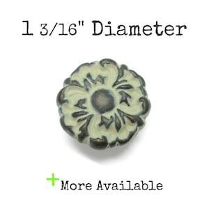 May include: A round knob with a floral relief pattern featuring a cream-colored surface and dark metallic accents. The knob has a 1 3/16 inch diameter. The text 1 3/16 inch Diameter and a green plus sign with the text More Available are visible.