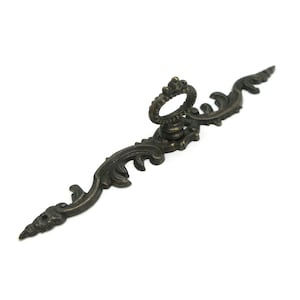 Peut inclure: Une poignée de tiroir en métal de couleur bronze présentant un design allongé et orné avec des motifs de feuilles en volutes et un anneau central. La quincaillerie comprend deux trous de montage aux extrémités effilées pour la fixation.