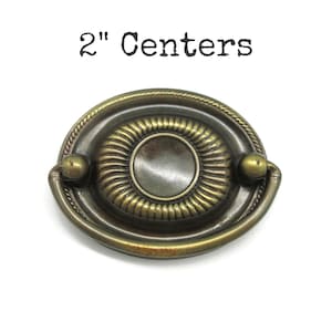 May include: An antique brass-toned drawer pull with an oval shape and a central circular design. The pull has a decorative rope-like border and two small ball-shaped accents. The text "2" Centers" is printed above the pull.