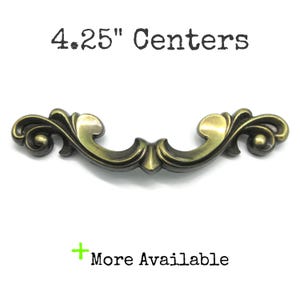 May include: An antique brass-coloured metal drawer pull with ornate, curved scrollwork detailing. The text 10.8 cm Centers is displayed above the handle, and a green plus sign with the text More Available is positioned below the hardware.