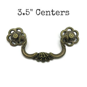 May include: An antique brass-toned metal drawer pull with a swinging handle and two circular floral-shaped mounting plates. The hardware features intricate scrollwork and shell-like detailing. The text 3.5" Centers is printed above the pull.