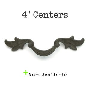 May include: A dark gray, decorative cabinet pull with a curved design and leaf-like accents. The pull has a 4-inch center-to-center measurement. Text at the top reads "4" Centers" and at the bottom "More Available".