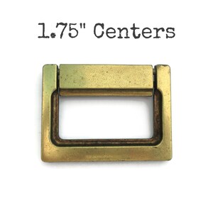 May include: A rectangular brass-toned metal drawer pull with a hinged drop handle. The hardware features a weathered finish and is labeled with the text 1.75" Centers, indicating the distance between the mounting points.