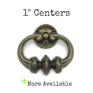 May include: Antique bronze-finished drawer pull with a decorative floral design. The pull has a ring handle and a 1-inch center-to-center measurement. The text "1" Centers" is above the pull, and "More Available" is below.