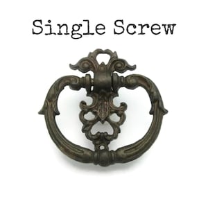 May include: Ornate, antique-style drawer pull with a dark bronze finish. The pull features a decorative oval shape with scrolling details and a central floral design. The text "Single Screw" is printed above the pull.