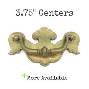 Peut inclure: Poignée de tiroir en laiton antique avec une poignée incurvée et une plaque arrière décorative. Le texte "3.75" Centers" est au-dessus de la poignée, et "More Available" est en dessous. La poignée mesure environ 9,5 cm de large.