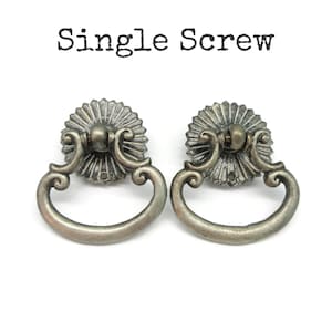 May include: Two antique brass-toned metal drawer pulls with a circular, sunburst-patterned backplate and a swinging drop handle. The text "Single Screw" is printed in black above the hardware, indicating the mounting style for these furniture pulls.
