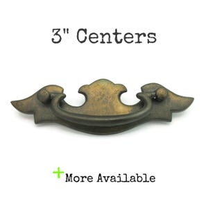 May include: Antique brass drawer pull with a curved handle and decorative ends. The text "3" Centers" is above the pull, and "More Available" is below. The pull measures 3 inches (7.6 cm) between the screw holes.