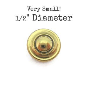 May include: A round, gold-colored metal button with a smooth, domed center and a raised circular rim. The text above the button reads: Very Small! 1/2" Diameter. The button has a polished, reflective finish and a simple, circular design.