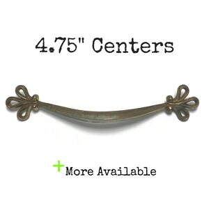 May include: A 4.75" center, antique gold, decorative cabinet pull with a curved shape and ornate, floral-shaped ends.