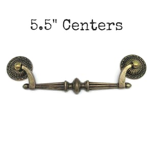 May include: A bronze-toned metal drawer pull with a central horizontal bar and two circular mounting plates featuring ribbed detailing. The hardware has a distressed finish and the text 5.5" Centers is displayed above the object.