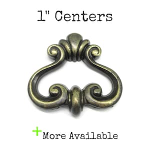 May include: A dark brass-toned metal drawer pull with a curved, ornate shape and a symmetrical design. The text 1" Centers is printed above the hardware, and the text + More Available is printed below the hardware on a plain white background.