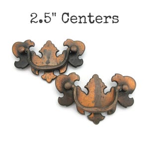 May include: Two antique-style drawer pulls with a distressed copper finish. Each pull features an ornate design with a curved handle and decorative end pieces. The text "2.5" Centers" is above the pulls.
