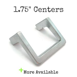 May include: A silver-colored cabinet pull with a square, open design. The text "1.75" Centers" is printed above the pull. The text "More Available" is printed below the pull.