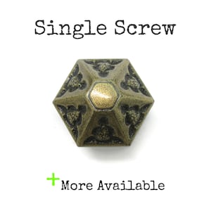 May include: A hexagonal brass-toned metal knob with a pyramid-shaped top and embossed floral patterns. The text "Single Screw" appears above the knob, and "+ More Available" is printed below in black text with a green plus sign.
