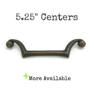 May include: Antique bronze-finished cabinet pull with a curved design and ball ends. The text "5.25" Centers" is printed above the pull, and "More Available" is printed below. This decorative hardware is suitable for drawers and cabinets.