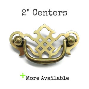 May include: A brass drawer pull with a decorative backplate featuring a lattice design and a swinging handle. The text 2" Centers is printed above the hardware, and the text + More Available is printed below the hardware on a white background.