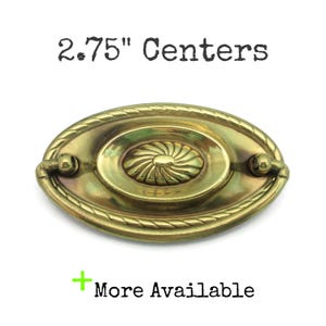May include: Oval brass drawer pull with a central floral design and rope-like edging. The text "2.75" Centers" is above the pull. The text "More Available" is below the pull. A decorative hardware piece.