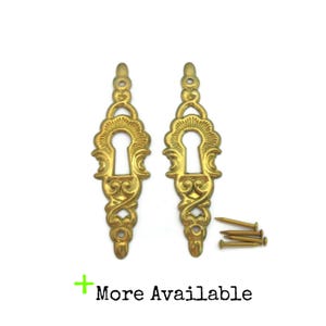 May include: Two ornate, gold-colored keyhole covers with decorative scrollwork and a teardrop shape. Includes a set of small brass nails. The text "More Available" is displayed in green.