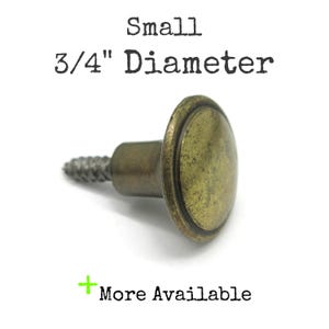 May include: A small brass-toned metal knob with a circular head and a threaded screw base. The text above reads Small 3/4" Diameter and the text below reads + More Available. The knob has a weathered, antique finish with a smooth, rounded top.