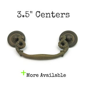 May include: Antique brass-toned drawer pull with a decorative design. The pull has a curved handle and two circular end pieces. The text "3.5" Centers" and "More Available" are printed on the image.