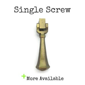 May include: A single brass cabinet pull with a square top and a pointed bottom. The pull is in a vintage style and has a slightly aged finish.
