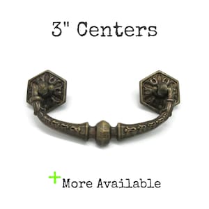 May include: Antique bronze drawer pull with a decorative floral design on the hexagonal end pieces. The pull measures 3 inches between the centers of the mounting holes. The text "3" Centers" is above the pull, and "More Available" is below.