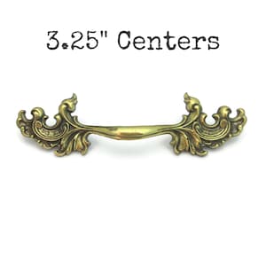 May include: Ornate brass drawer pull with a curved handle and decorative floral accents. The text "3.25" Centers" is above the pull. This vintage-style hardware is perfect for adding a touch of elegance to furniture.