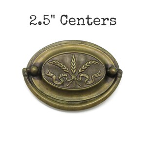 May include: An oval-shaped brass drawer pull with a dark antique finish, featuring a central embossed wheat stalk and ribbon design. The hardware includes a swinging bail handle and is labelled with the text 2.5" Centers above the object.
