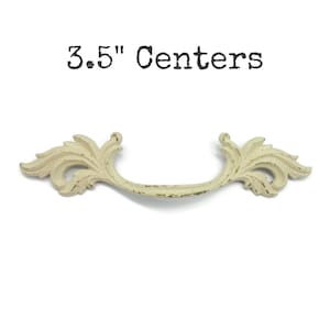 May include: A cream-colored drawer pull with a distressed finish and ornate leaf-like detailing on both ends. The handle features a curved design and is labeled with the text 3.5" Centers, indicating the distance between the mounting points.