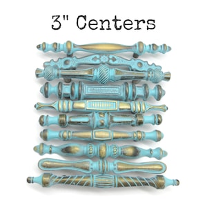 May include: A collection of ten drawer pulls with 3" centers, featuring a distressed turquoise blue finish with exposed antique gold metal highlights. Each handle displays a unique decorative shape, including ribbed, smooth, and textured patterns.