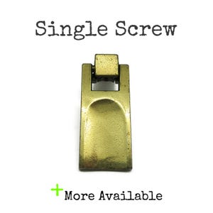 May include: A brass-toned metal furniture pull with a rectangular base and a hinged top section. The text "Single Screw" appears at the top, and the text "+ More Available" is positioned at the bottom of the image.