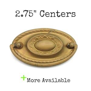May include: An antique brass oval drawer pull with a decorative rope-like border and an oval center. The text "2.75" Centers" is above the pull. The text "More Available" is below the pull.