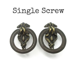 May include: Two antique brass-toned metal ring pulls featuring ornate, textured mounting plates with a central oval detail. The text "Single Screw" is printed in black above the hardware. These circular drawer pulls have a dark, weathered finish.