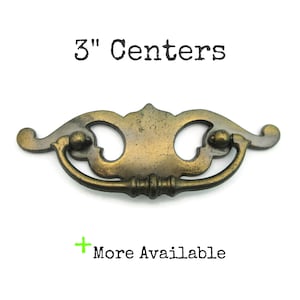 May include: Antique brass drawer pull with a decorative design. The pull has a curved handle and measures 3 inches between the centers of the mounting holes. The text "3" Centers" is above the pull, and "More Available" is below.
