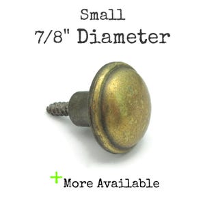 May include: A small, antique-style brass knob with a 7/8" diameter. The knob has a rounded, domed shape and a threaded screw for attachment. The text "Small 7/8" Diameter" is printed above the knob. The text "More Available" is printed below the knob.
