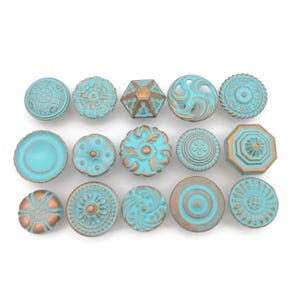 May include: A collection of fifteen decorative cabinet knobs in various shapes and designs. The knobs feature a distressed turquoise finish with bronze accents. The designs include floral, geometric, and circular patterns, offering a vintage aesthetic.