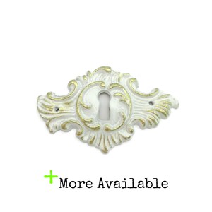 May include: White and gold ornate keyhole cover with a decorative design. The keyhole cover has a central keyhole and two screw holes. The text "More Available" is below the keyhole cover.