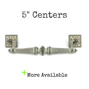 May include: Antique-style cabinet pull with a distressed finish. The pull measures 5 inches (12.7 cm) between centers. Square backplates and a decorative center section add to the vintage aesthetic. The text "5" Centers" and "More Available" are visible.