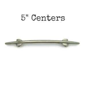 May include: A silver-toned cabinet pull with a curved design. The pull has a 5-inch (12.7 cm) center-to-center measurement and is set against a white background. The text "5" Centers" is above the pull.