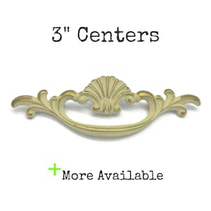 May include: A cream-colored metal drawer pull with gold-toned highlights and a central shell motif. The hardware features a curved handle design and is labeled with the text 3" Centers and More Available.