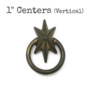 May include: Antique brass drawer pull with a starburst design and a circular handle. The text "1" Centers (Vertical)" is above the pull. The pull measures 2.54 cm in length.