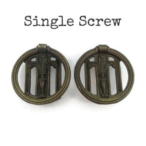 May include: Two circular bronze-toned metal drawer pulls featuring a central urn and leaf motif within a ring frame. The text "Single Screw" is printed above the hardware. These decorative pulls have a dark, aged finish and a hinged ring design.
