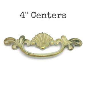 May include: Antique brass drawer pull with a shell-shaped center and ornate floral details. The pull measures 4 inches between the centers of the mounting holes. The text "4" Centers" is above the pull.