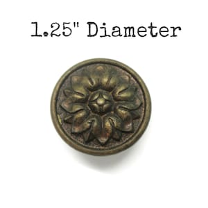 May include: A round, antique brass-coloured metal knob featuring a raised, symmetrical floral pattern with multiple petals radiating from a central point. The text 1.25" Diameter is printed above the knob, indicating its specific size measurement.