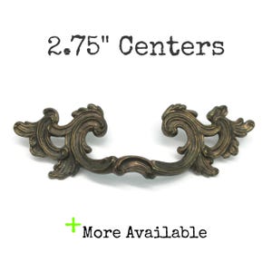 May include: An ornate bronze-toned metal drawer pull with scrolling leaf-like details and a curved handle. The text 2.75" Centers is displayed above the hardware, and the text + More Available is positioned below the object.