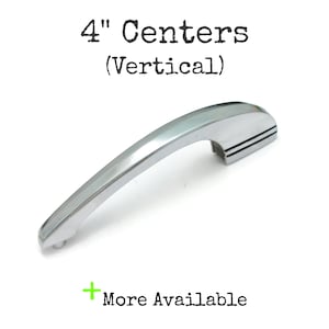May include: A polished chrome cabinet handle with a curved, elongated design. The text above reads 4" Centers (Vertical) and the text below reads + More Available. The handle features a smooth, reflective metallic finish and a sturdy mounting base.