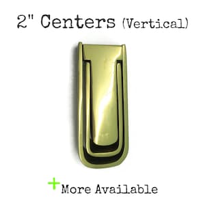 May include: A polished brass vertical drop pull handle with a rectangular frame and a U-shaped swinging handle. The text 2" Centers (Vertical) and + More Available is printed in black above and below the hardware piece respectively.