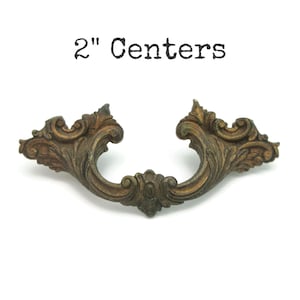 May include: Ornate antique bronze drawer pull with a curved design. The pull has a floral and scrollwork pattern and measures 2 inches between the centers of the mounting holes. The text "2" Centers" is above the pull.
