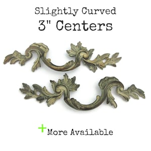 May include: Two antique-style drawer pulls with a slightly curved design. The pulls are a bronze colour with ornate leaf and scroll details. The text "Slightly Curved 3" Centers" is at the top. The text "More Available" is at the bottom.